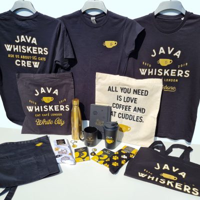 Java Whiskers Products - 3 Black T-shirts with Java Whiskers logos on, a black bag with the logo on, a cream bag with a slogan on. a gold bottle with paw print design. Notebook with paws for thought and paw design. a Mug and travel mug with logo on, an apron with the java cup logo. socks with illustrations and paw prints. Pin badges and keyring with logos and paw print designs. and another black bag with logo.